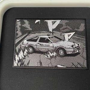 Pode incluir: Um remendo bordado em preto e branco que representa um carro branco dirigindo em uma estrada com um fundo cinza. O carro tem a palavra "REBEL" escrita no lado. O remendo é cercado por uma borda preta.