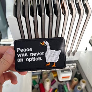 Op de afbeelding: Zwart rechthoekig embleem met de tekst "Peace was never an option." en een witte ganzenillustratie met oranje voeten en snavel. Het embleem wordt voor een borduurmachine met garenklosjes gehouden.