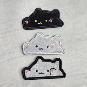 Puede incluir: Tres parches bordados de gato en negro, gris y blanco. Cada parche presenta una cara de gato con una boca sonriente, huellas de patas rosas y ojos amarillos o negros. Los parches están delineados en negro o blanco.