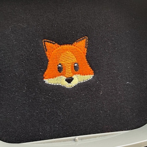 Puede incluir: Diseño bordado de la cara de un zorro sobre una tela negra. El zorro tiene pelaje naranja, detalles blancos alrededor de los ojos y la nariz, y ojos negros.