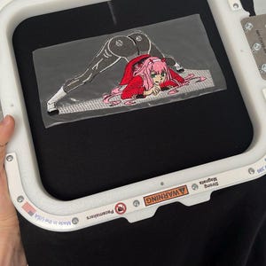 Puede incluir: Parche bordado con un personaje de anime con cabello rosado y un atuendo rojo. El personaje está en posición de rodillas con la espalda arqueada. El parche está sobre un fondo de tela negro.