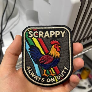 May include: Embroidered patch with a black border featuring a colorful rooster design. The patch has the text "SCRAPPY" at the top and "ALWAYS ON DUTY" around the bottom edge. The rooster is in shades of red, yellow, blue, and green.