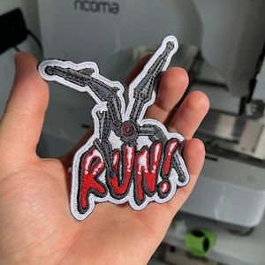May include: Embroidered patch featuring a robotic spider design with the word "RUN!!" in red and white. The patch has a black border and is held in a hand.