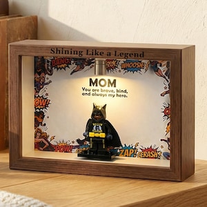 Puede incluir: Una caja de sombra de madera con el texto "Shining Like a Legend" y "MOM". En el interior hay una figura de superhéroe y gráficos de estilo cómic. El texto dice "You are brave, kind, and always my hero."