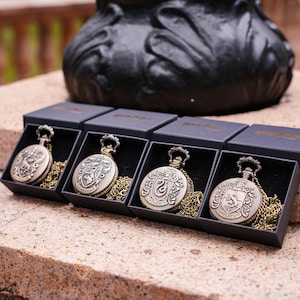 May include: Four antique-style pocket watches in black boxes, each with a different house crest from a fantasy series. The watches are bronze-colored with matching chains. The boxes are on a stone surface.