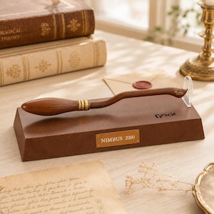 May include: A brown wooden broomstick replica featuring gold-colored bands near the bristles and the text Nimbus 2000 engraved on the handle. The broom rests on a brown rectangular display base with a gold plaque reading NIMBUS 2000.