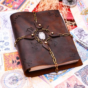 May include: A brown leather journal with a crystal and a chain wrapped around it. The journal has various symbols and writing on the cover.
