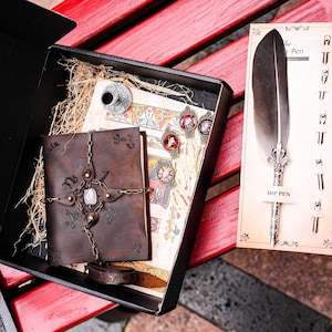May include: A brown leather journal with a metal chain and a crystal charm, a quill pen with a feather, and a small inkwell. The journal is open to reveal a page with a decorative design. The quill pen is on a card that says "Dip Pen".
