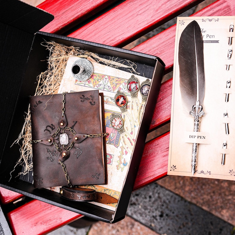 May include: A brown leather journal with a metal chain and a crystal charm, a quill pen with a feather, and a small inkwell. The journal is open to reveal a page with a decorative design. The quill pen is on a card that says "Dip Pen".