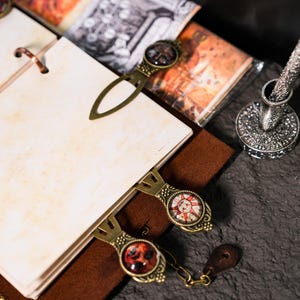May include: A brown leather bound journal with blank pages and three brass bookmarks with decorative glass insets. The bookmarks feature images of a sun, a skull, and a cross.