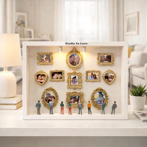 Puede incluir: Una caja de sombra blanca con m&uacute;ltiples fotos de parejas enmarcadas en oro. Las fotos tienen varias formas, incluyendo rectangulares y en forma de coraz&oacute;n. El texto "Rushe In Love" est&aacute; encima de las fotos. Figuras en miniatura est&aacute;n debajo.
