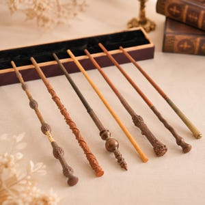 May include: A collection of six distinct wands arranged in a rectangular display box. The wands feature varied designs including textured wood-like finishes, smooth tapered shafts, and metallic accents in shades of brown, tan, and bronze.