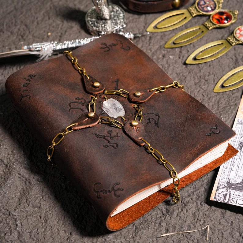 May include: A brown leather journal with a chain and a clear crystal. The journal has a decorative design on the cover and is closed.