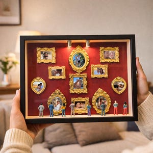 Puede incluir: Una caja de sombra con marco negro y fondo rojo, que muestra m&uacute;ltiples marcos de fotos dorados y ornamentados. Los marcos contienen varias fotograf&iacute;as. Figuras en miniatura est&aacute;n posicionadas en la parte inferior de la caja.