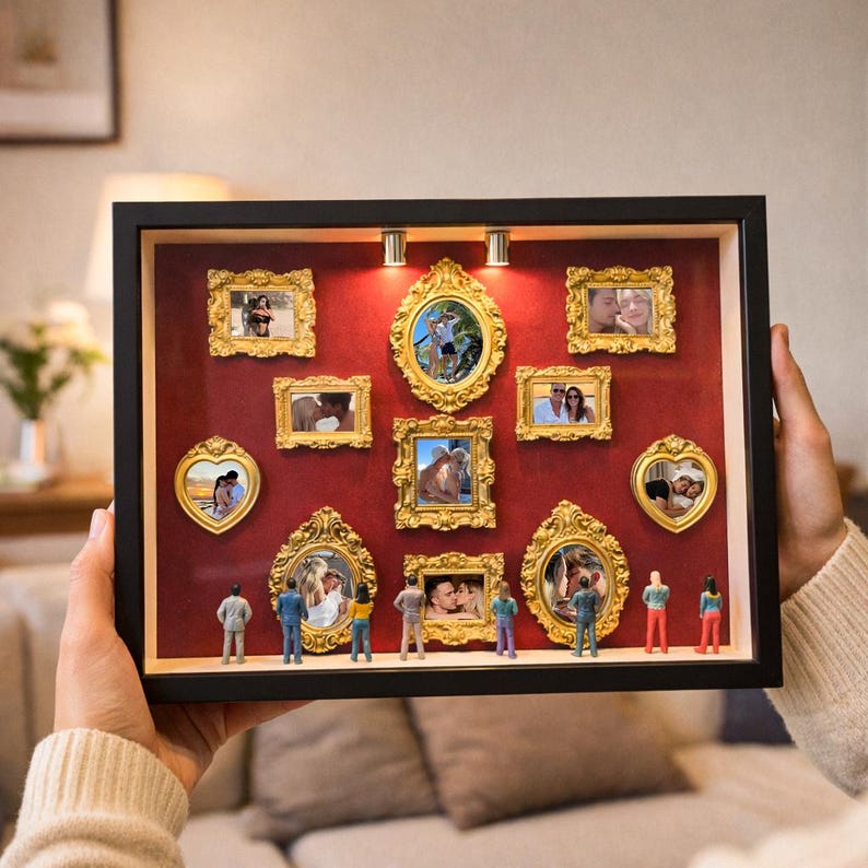 Puede incluir: Una caja de sombra con marco negro y fondo rojo, que muestra m&uacute;ltiples marcos de fotos dorados y ornamentados. Los marcos contienen varias fotograf&iacute;as. Figuras en miniatura est&aacute;n posicionadas en la parte inferior de la caja.