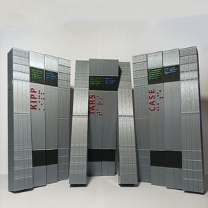 Può includere: Quattro scatole rettangolari di colore argento con accenti neri e testo rosso. Le scatole sono etichettate "KIPP", "TARS", "CASE" e "..." Le scatole sono disposte in fila da sinistra a destra.