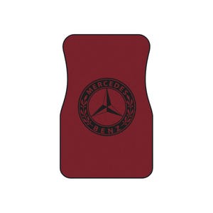 May include: A burgundy car floor mat featuring the Mercedes-Benz logo in black. The logo is a three-pointed star within a circle, surrounded by a laurel wreath, with the words "Mercedes Benz" encircling it.