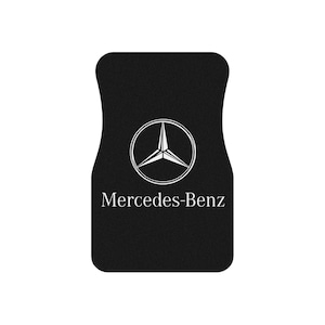 May include: Black car floor mat featuring the Mercedes-Benz logo in white. The logo is a three-pointed star inside a circle. The words "Mercedes-Benz" are printed below the logo in a clean, sans-serif font. The mat has a contoured shape.
