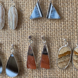 May include: A set of eight silver earrings with various stone designs. The stones are in shades of brown, white, and blue. The earrings are displayed on a brown burlap background.