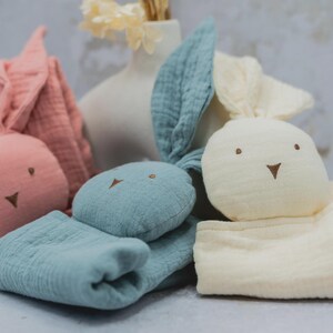 Peut inclure: Trois jouets en forme de lapin aux couleurs pastel. Un rose, un bleu et un crème. Chacun a des traits de visage brodés et des oreilles tombantes. Les jouets sont en tissu texturé, probablement de la mousseline, et sont conçus pour les bébés.