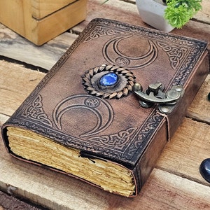 May include: A brown leather journal with a Celtic knot design and three crescent moons embossed on the cover. The journal has a metal clasp and a blue gemstone set in a metal embellishment.