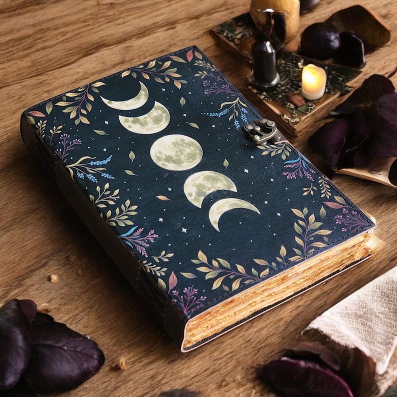 May include: A dark blue journal with a moon phases design and floral accents. The cover features a series of moon phases, surrounded by leaves and flowers. The journal has a metal clasp and aged-looking pages, suggesting a vintage or mystical theme.