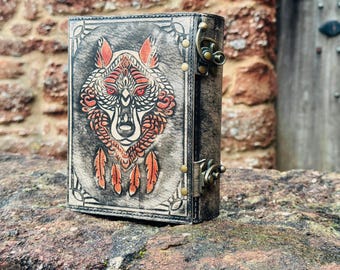 Pode incluir: Um diário de couro preto com um design de lobo estampado na capa frontal. O lobo é branco com detalhes vermelhos e tem penas saindo de sua cabeça. O diário tem duas travas de latão.