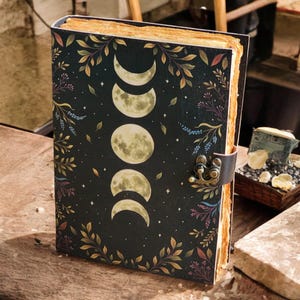 May include: A hardcover journal with a black cover featuring a moon phases design and botanical accents. The journal has a leather strap closure and aged paper pages. The cover design includes crescent moons and full moons, with floral and leaf details.