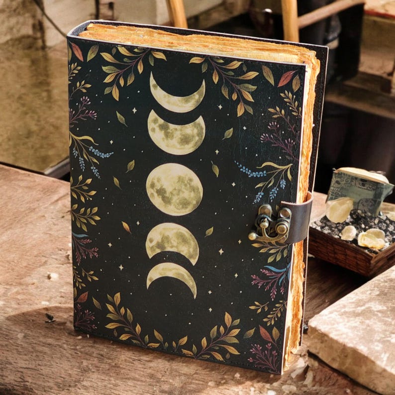 May include: A hardcover journal with a black cover featuring a moon phases design and botanical accents. The journal has a leather strap closure and aged paper pages. The cover design includes crescent moons and full moons, with floral and leaf details.