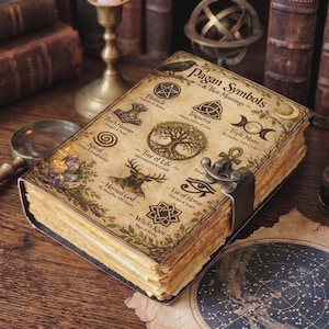 Peut inclure: Un livre rectangulaire avec une couverture en parchemin vieilli intitulée Pagan Symbols & Their Meanings. La couverture présente des illustrations d'un pentacle, d'une triquetra, d'une triple lune, d'une spirale, d'un arbre de vie et d'un nœud de sorcière.