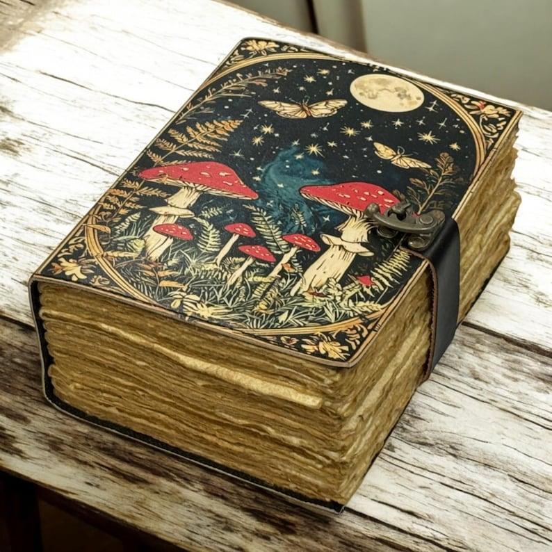 May include: A decorative journal with a black leather strap closure. The cover features a whimsical illustration of red mushrooms, butterflies, and a full moon against a starry night sky. The pages have a vintage, aged appearance.