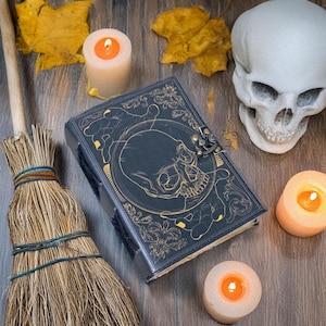 Peut inclure: Un journal en cuir noir avec un motif de crâne doré et un fermoir en métal. Le journal est entouré de bougies, d'un balai et d'un crâne blanc.