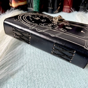 Puede incluir: Un diario de cuero negro con un cierre plateado y un dise&ntilde;o detallado de una puerta con una cita en un c&iacute;rculo a su alrededor. El diario tiene costuras negras a lo largo del lomo.