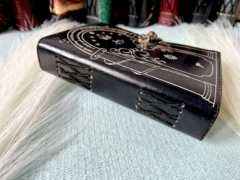 Puede incluir: Un diario de cuero negro con un cierre plateado y un dise&ntilde;o detallado de una puerta con una cita en un c&iacute;rculo a su alrededor. El diario tiene costuras negras a lo largo del lomo.