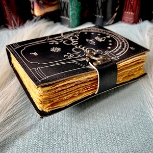 Puede incluir: Un diario encuadernado en cuero negro con un dise&ntilde;o blanco de un &aacute;rbol y una puerta. El diario tiene un cierre de metal y est&aacute; cerrado.