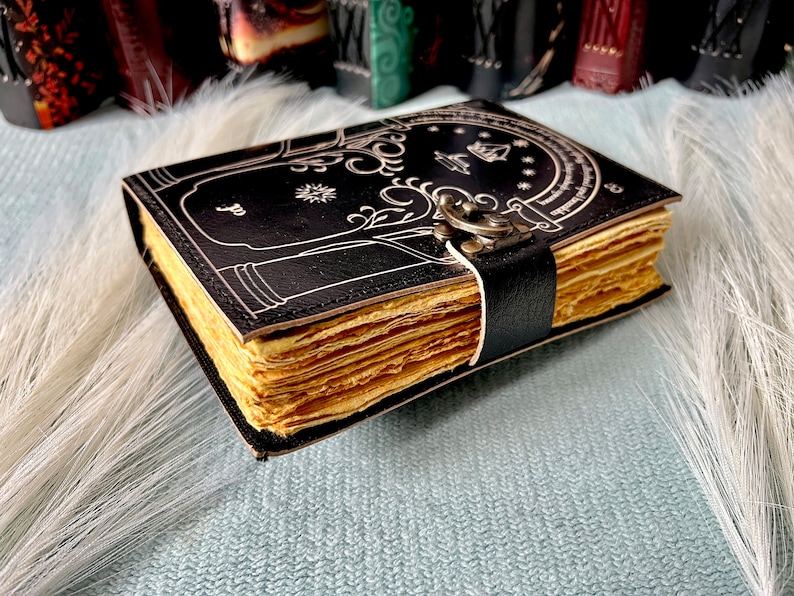 Puede incluir: Un diario encuadernado en cuero negro con un dise&ntilde;o blanco de un &aacute;rbol y una puerta. El diario tiene un cierre de metal y est&aacute; cerrado.