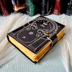 Puede incluir: Un diario de cuero negro con una ilustraci&oacute;n detallada de un &aacute;rbol y una puerta, con un cierre de metal. El diario tiene un borde dorado y est&aacute; cerrado.