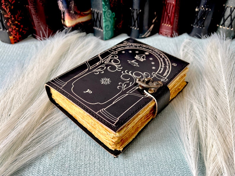 Puede incluir: Un diario de cuero negro con una ilustraci&oacute;n detallada de un &aacute;rbol y una puerta, con un cierre de metal. El diario tiene un borde dorado y est&aacute; cerrado.