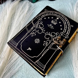 Puede incluir: Diario de cuero negro con un dise&ntilde;o blanco de un &aacute;rbol y una puerta con una cita en letras blancas alrededor del borde. El diario tiene un cierre de metal.