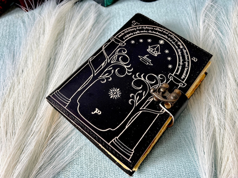 Puede incluir: Diario de cuero negro con un dise&ntilde;o blanco de un &aacute;rbol y una puerta con una cita en letras blancas alrededor del borde. El diario tiene un cierre de metal.