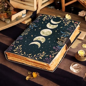 May include: A vintage-style journal with a dark blue cover featuring a moon phase design, stars, and botanical illustrations. The journal has a leather strap closure and aged, yellowed pages, suggesting a well-loved and cherished item. The overall aesthetic is reminiscent of a mystical or fantasy theme.