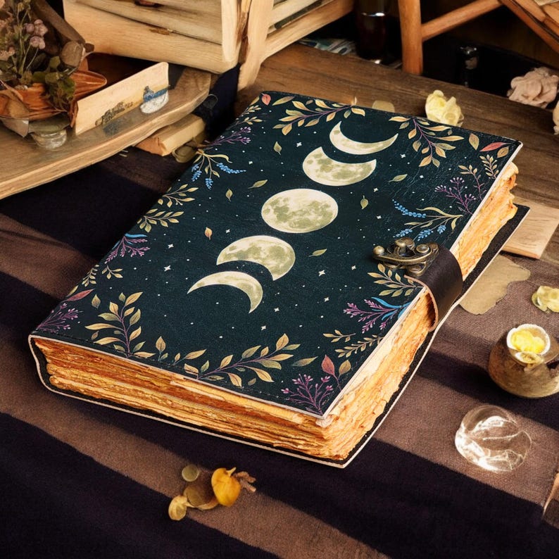 May include: A vintage-style journal with a dark blue cover featuring a moon phase design, stars, and botanical illustrations. The journal has a leather strap closure and aged, yellowed pages, suggesting a well-loved and cherished item. The overall aesthetic is reminiscent of a mystical or fantasy theme.