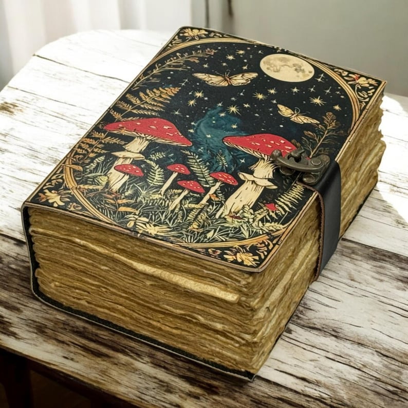 May include: A vintage-style journal with a detailed cover illustration. The cover features red mushrooms, butterflies, and a full moon against a starry night sky. The journal has a black leather strap closure and aged, yellowed pages.