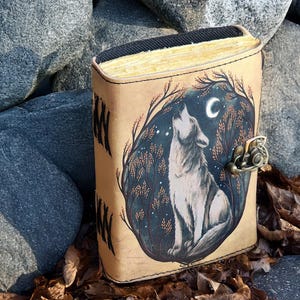 May include: A tan leather journal with a detailed illustration of a howling wolf under a crescent moon. The artwork is set within a dark forest scene. The journal has a metal clasp and visible, aged paper edges.