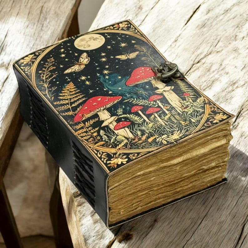 May include: A leather-bound journal with a whimsical design. The cover features a night sky scene with mushrooms, butterflies, and a full moon. The journal has a metal clasp and aged-looking pages.