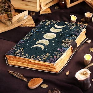 May include: A dark blue journal with a celestial design featuring moon phases and stars. The cover is adorned with botanical illustrations in shades of green, purple, and gold. The journal has a leather strap closure and aged-looking pages.