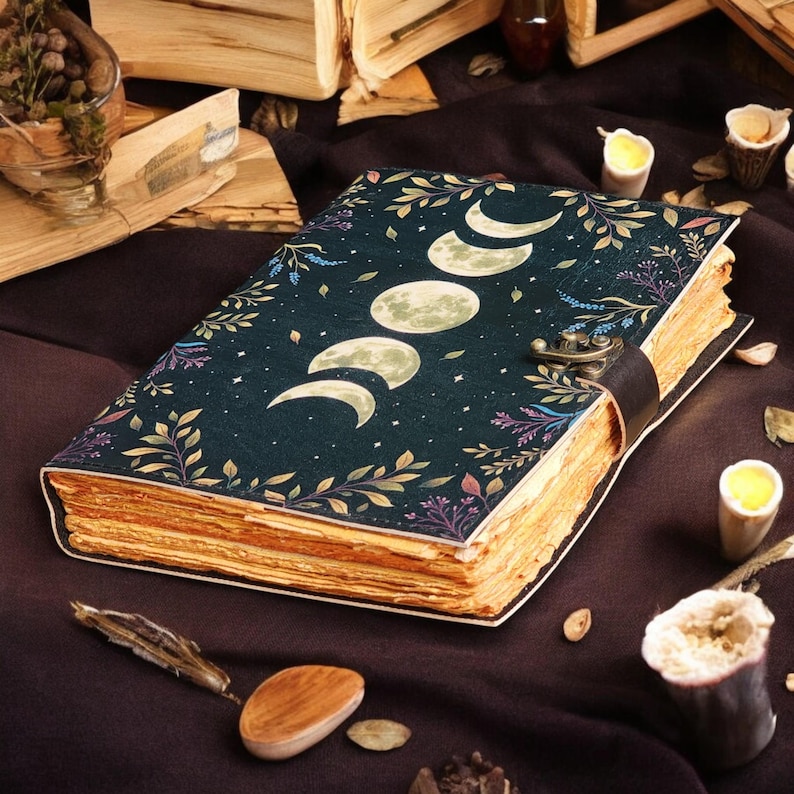 May include: A dark blue journal with a celestial design featuring moon phases and stars. The cover is adorned with botanical illustrations in shades of green, purple, and gold. The journal has a leather strap closure and aged-looking pages.