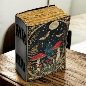 May include: A leather-bound journal with a whimsical mushroom and butterfly design. The cover features a dark blue background with red mushrooms, butterflies, and a full moon. The journal has a metal clasp and stitched spine.