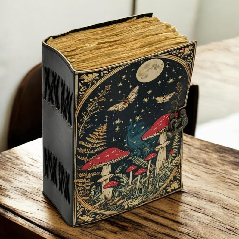 May include: A leather-bound journal with a whimsical mushroom and butterfly design. The cover features a dark blue background with red mushrooms, butterflies, and a full moon. The journal has a metal clasp and stitched spine.