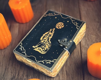 Pode incluir: Um diário encadernado em couro preto com um desenho de lobo dourado uivando para a lua. O diário tem um fecho de fivela e está rodeado por velas laranja.